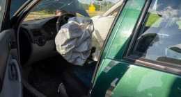 Egy zöld színű, nyitott első ajtóval rendelkező autó vezető oldalán kinyílt légzsákok és megrepedt szélvédő jelzi, hogy balesetet szenvedett. A motorháztető felhúzva, és kintről süt a nap.