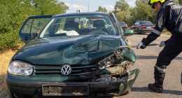Egy egyenruhás tűzoltó szerszámot használ egy út szélén parkoló zöld színű, súlyosan sérült Volkswagen személygépkocsin. Az autó ajtaja nyitva van, a háttérben pedig más járművek sorakoznak az úton.