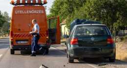 Egy rendőr megvizsgál egy megrongált zöld VW Golfot, amely az út szélén parkol egy narancssárga színű, Útellenőrzés feliratú útszolgálati kisteherautó mellett. Az úton autóroncsok hevernek, a háttérben fák és házak.