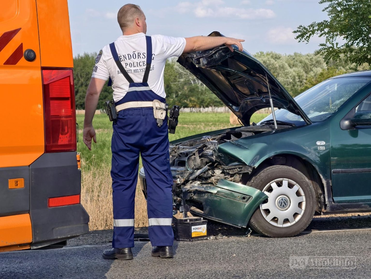 Egy rendőr áll egy narancssárga jármű mellett, és egy zöld autó felé mutat, amelynek az eleje összeroncsolódott, a motorháztetője pedig nyitva van egy ütközés után, egy fűvel és fákkal szegélyezett úton.