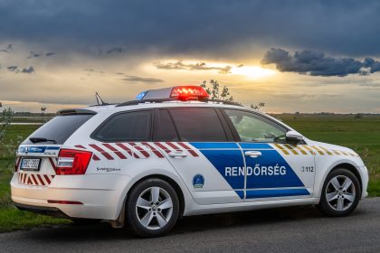 Egy magyar rendőrautó kék és piros lámpákkal az út szélén parkol naplementekor, a háttérben mezők és felhős égbolt.